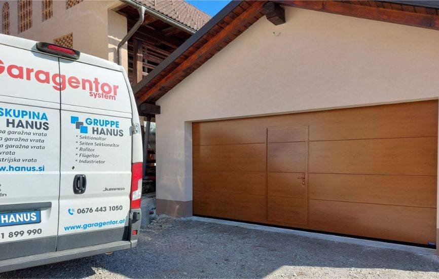 Parkiran kombi podjetja Hanus pred sekcijskimi garaznimi vrati z imitacijo lesa.